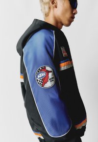 Crna i plava jakna s kapuljačom, s narančastim i bijelim prugama. Uključuje kružni KTM Racing Team pečat na lijevom rukavu.