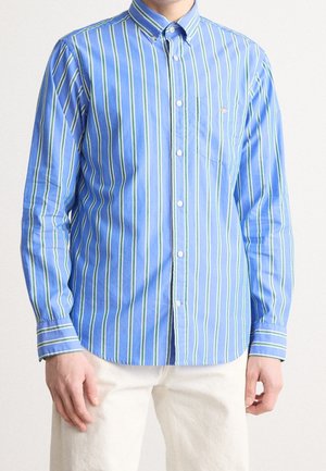 Muž v modré košili na knoflíky s vertikálními zelenými a bílými pruhy a bílých kalhotách, stojící před jednoduchým pozadím.