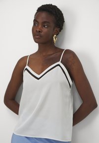Hvit sateng camisole med tynne stropper, med en svart V-formet utringning. Jevn tekstur, løs passform og subtil glans.