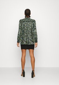 Blusa nera trasparente a fiori con maniche lunghe, caratterizzata da un motivo botanico verde e blu, abbinata a una gonna nera aderente e tacchi.