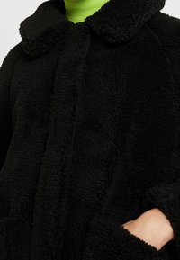 Czarny, pluszowy, oversized płaszcz z kołnierzem i zapięciem na guziki z przodu. Posiada miękki, teksturowany materiał oraz duże kieszenie boczne.