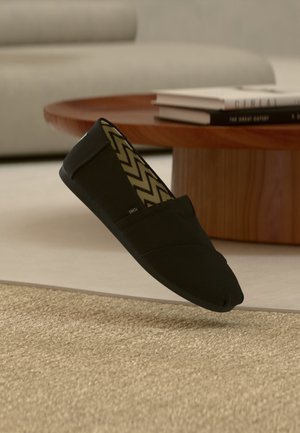 Chaussure noire en toile sans lacets avec une semelle intérieure à motif chevron flottant au-dessus d'un tapis beige, près d'une table ronde en bois avec des livres empilés.