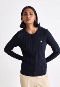 Donna che indossa un cardigan blu navy a trecce con bottoni e pantaloni beige, in piedi con le mani dietro la schiena contro uno sfondo bianco semplice.