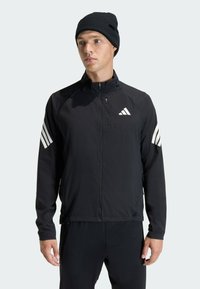Black lightweight zip jacket with white Adidas logo and three-striped accents on shoulders, paired with a black knit beanie.
