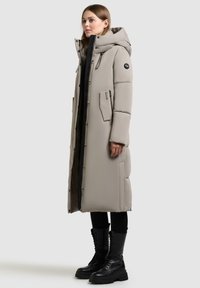 Lang beige puffermantel med hætte, to sidelommer og lynlås foran. Parret med sorte kampstøvler for et kontrastfuldt look.
