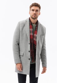 Cappotto grigio in lana con rever a punta, chiusura con bottoni e tasche laterali, abbinato a una sciarpa a quadri rossi e neri. Sotto, un maglione con texture.