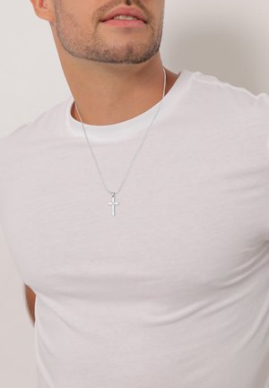 Silver cross pendant necklace on a thin chain, worn over a fitted white t-shirt. The pendant features a smooth, polished surface.