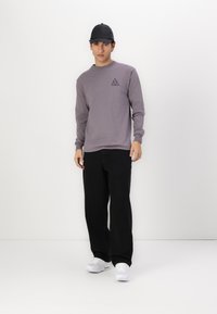 Grå longsleeve skjorte med et lite svart trekantet logo og svarte bukser, båret med hvite sneakers og en svart caps. Enkelt bakgrunn.