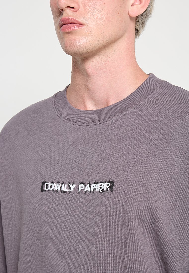 Grauer Kapuzenpullover mit geripptem Halsausschnitt und einem auffälligen Logo in Schwarz und Weiß, das über die Brust gestickt ist. Weicher Stoff.