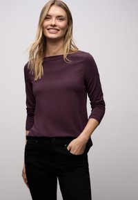 Fioletowa bluzka z długim rękawem i dekoltem w łódkę, wykonana z gładkiej tkaniny. Połączona z czarnymi spodniami, prezentująca prostą, dopasowaną sylwetkę.
