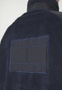 Granatowa polarowa kurtka z teksturowaną powierzchnią i naszywką z napisem "TOMMY JEANS" w kontrastującym niebieskim kolorze.