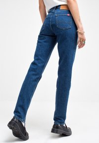 Jeans de mezclilla azul con corte recto, que cuentan con dos bolsillos traseros y una etiqueta de cuero en la cintura. Combinados con zapatos de plataforma negros.