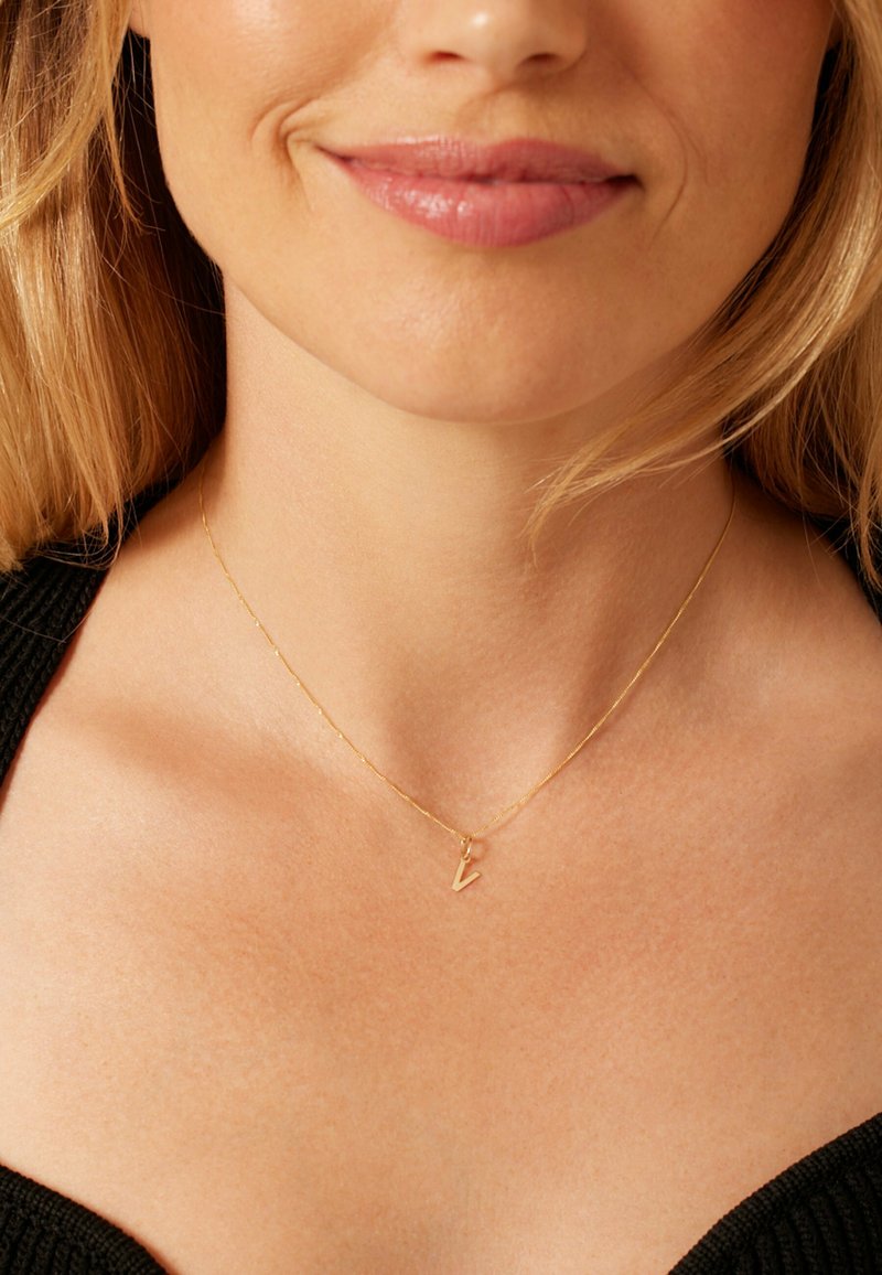 Gouden ketting met een fijne schakelketting en een kleine 'V' hanger. De achtergrond is een gestructureerd zwart shirt.