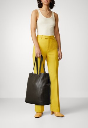 Femme portant un débardeur blanc, un pantalon taille haute jaune, des chaussures à talons beiges, tenant un grand sac cabas noir texturé sur son côté gauche.