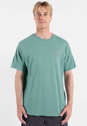 T-shirt en coton vert avec col rond et manches courtes, arborant une broderie florale beige sur le côté gauche de la poitrine.