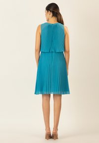 Türkises, plissiertes Kleid mit einem geschichteten Rückendesign, ärmellosem Schnitt und einem fließenden Material, kombiniert mit transparenten Absätzen.