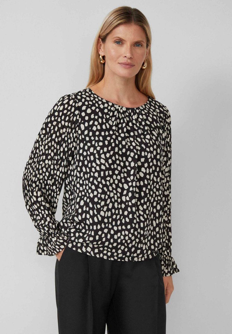 Schwarze Bluse mit weißem abstraktem Muster, runder Ausschnitt, lange Ärmel und Raffung an den Schultern und Bündchen. Glattes Material.