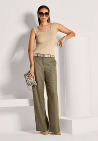 Beige ärmelloses Top, grüne strukturierte weite Hose, pinker Gürtel mit runder Schnalle, silberne Handtasche mit Kette, übergroße dunkle Sonnenbrille.