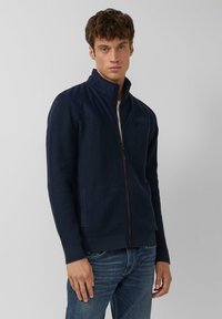 Veste zippée bleu marine avec un design en tricot texturé, un panneau supérieur lisse, des poches latérales et des poignets élastiques. Portée par-dessus une chemise blanche et un jean.