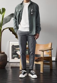 Chemise verte boutonnée portée sur un pull en maille torsadée crème, pantalon gris ajusté et baskets noires à semelles blanches. Cadre intérieur avec des plantes.