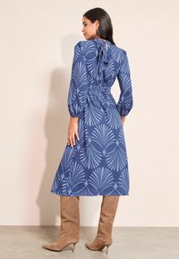 Zila kleita ar palmu lapu rakstu, pieguloša josta un garām piedurknēm. Apvienota ar krēmbaltām augstpapēžu kurpēm līdz augšstilbiem. Gluda auduma un strukturēts dizains.