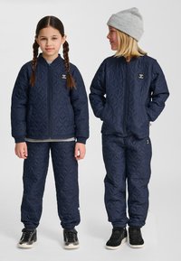 Dwoje dzieci ubranych w pasujące granatowe pikowane kurtki i spodnie, jedno z warkoczykami, drugie w szarej czapce, stojące obok siebie.