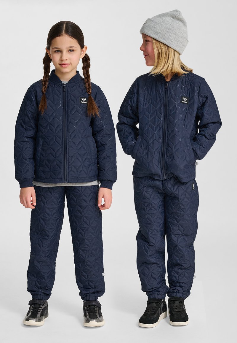 Dwoje dzieci ubranych w pasujące granatowe pikowane kurtki i spodnie, jedno z warkoczykami, drugie w szarej czapce, stojące obok siebie.