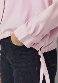 Osoba ubrana w jasnoróżową koszulę z długim rękawem z zawiązanym dołem oraz ciemnoniebieskie dżinsy, z ręką schowaną w kieszeni.