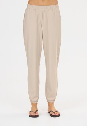 Beige elastiske joggingbukser med sidelommer båret af en person med sorte flip-flops på en hvid baggrund.