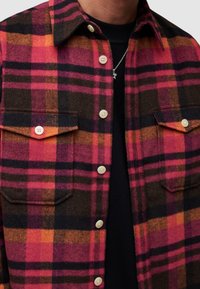 Koszula z wełny w kratę w kolorze różowym, pomarańczowym, czarnym i brązowym. Posiada dwie kieszenie na piersi, zapięcie na guziki oraz kołnierzyk. Miękka tekstura.