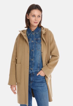 Beige wool coat with a hood, front pockets, and drawstring. Layered over a blue denim jacket, featuring a buttoned placket and collar.