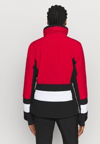 Persona con el cabello trenzado que lleva una chaqueta bloqueada en colores rojo, negro y blanco, vista desde atrás contra un fondo liso.