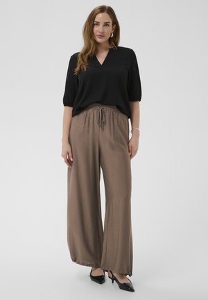 Naine, kes kannab musta V-kaelusega pluusi küünarvarrepikkuste varrukatega, pruune laiade säärtega säästvaid pükse ja musti teravate otsaga slingback-kingi ning hõbedast käevõru.