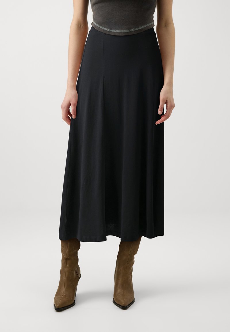 Femme portant une jupe midi noire, un haut gris ajusté et des bottes en daim marron montantes jusqu'aux genoux, debout devant un fond clair uni.