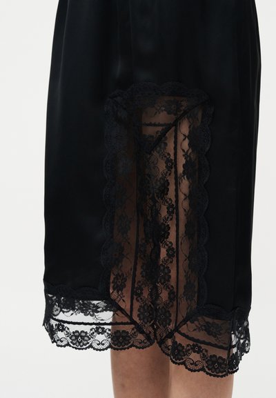 Μαύρη σατέν φούστα με διάφανο μαύρο δαντελένιο πάνελ με λουλούδια και σκάλιστο δαντελένιο τελείωμα, που αφήνει να φανεί μέρος των γυμνών ποδιών μέσα από τη δαντέλα.