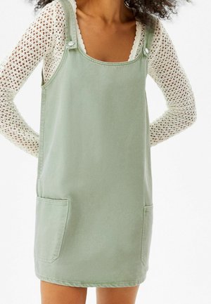 Femme portant une robe salopette sans manches vert clair par-dessus un haut en maille blanche à manches longues, debout les mains derrière le dos.