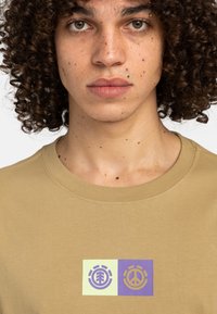 Beiges Rundhals-T-Shirt mit zwei grafischen Drucken: einem grünen mit einem Baumsymbol und einem lila mit einem Friedenssymbol. Weiches Baumwollmaterial.