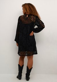 Rochie neagră din dantelă cu mâneci lungi, model floral și detaliu de tăietură în formă de cheie la spate, asortată cu cizme cowboy negre. Textura este diafană și stratificată.