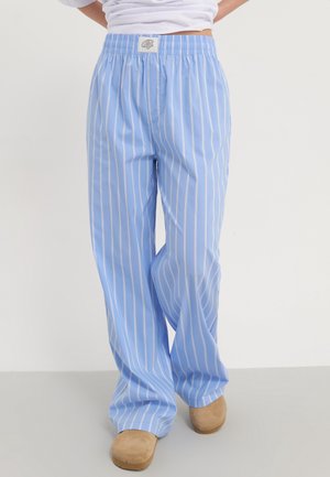 Personne portant un pantalon bleu clair à rayures blanches verticales et des chaussures beiges à enfiler, debout devant un fond uni.