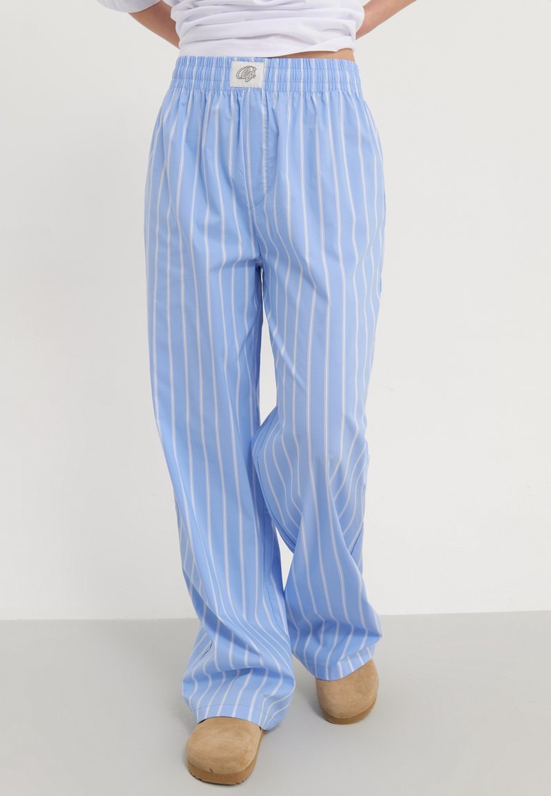 Personne portant un pantalon bleu clair à rayures blanches verticales et des chaussures beiges à enfiler, debout devant un fond uni.