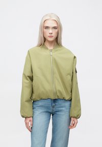 Oliven grønn bomberjakke med glidelås foran, elastiske mansjetter og nederkant, samt sidelommer. Kombinert med lyseblå denimjeans.