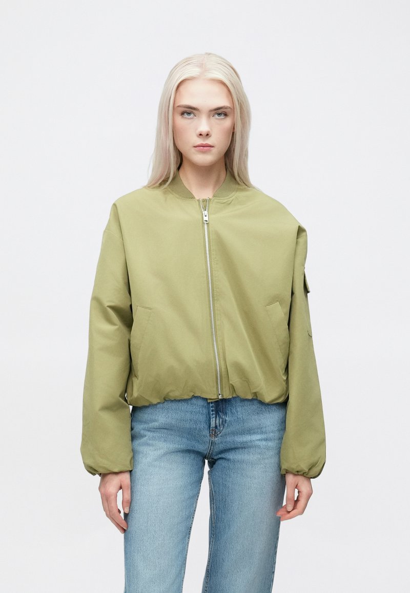 Oliven grønn bomberjakke med glidelås foran, elastiske mansjetter og nederkant, samt sidelommer. Kombinert med lyseblå denimjeans.