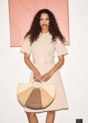 Kvinde med krøllet hår iført en beige knap-kjole, der holder en halvcirkelformet stråtaske, stående mod en hvid væg med lyserød kunst.