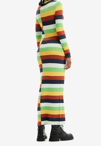 Femme portant une longue robe rayée multicolore côtelée et des bottes noires à lacets avec des motifs floraux, se tenant devant un fond blanc.