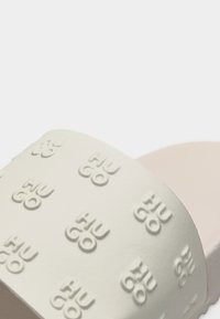 Gros plan sur la sangle beige d'une sandale slide embossée de logos "HUGO" répétés en lettres en relief ton sur ton.