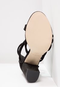 Sandale noire à talon haut avec plusieurs brides, vue du côté de la semelle, affichant une semelle beige lisse et un bout de talon carré.