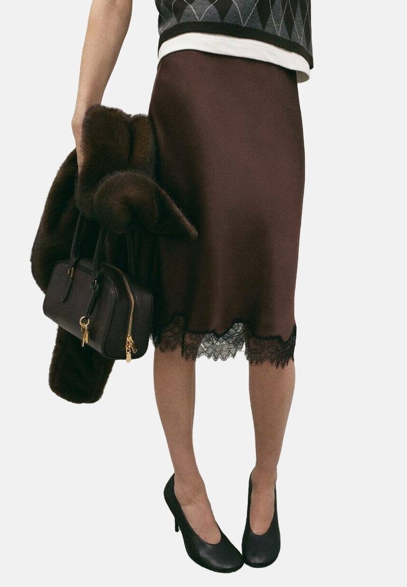 Femme portant une jupe marron avec une bordure en dentelle, tenant un manteau en fourrure foncé et un sac à main noir, et portant des talons hauts noirs.