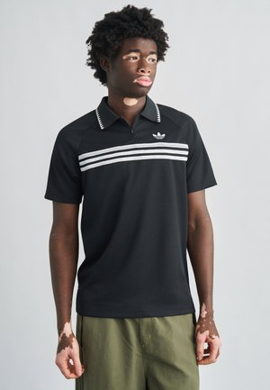 Czarna koszulka polo z zamkiem błyskawicznym, białymi poziomymi paskami i kołnierzykiem. Na piersi znajduje się logo Adidas. W zestawie z oliwkowymi szortami.