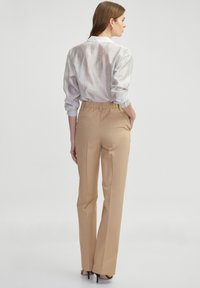Pantalones beige con ajuste entallado, que cuentan con bolsillos laterales y una cinturilla. El modelo lleva una camisa de manga larga de rayas claras.