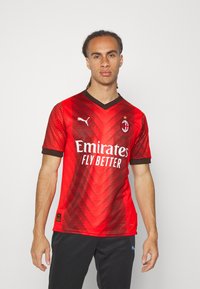 Maglia da calcio a strisce rosse e nere con scollatura a V, caratterizzata da un logo prominente, testo dello sponsor e un motivo testurizzato. Maniche corte.
